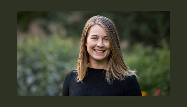 Dr. Maaike Canrinus stands in front of foliage, smiling at the camera.